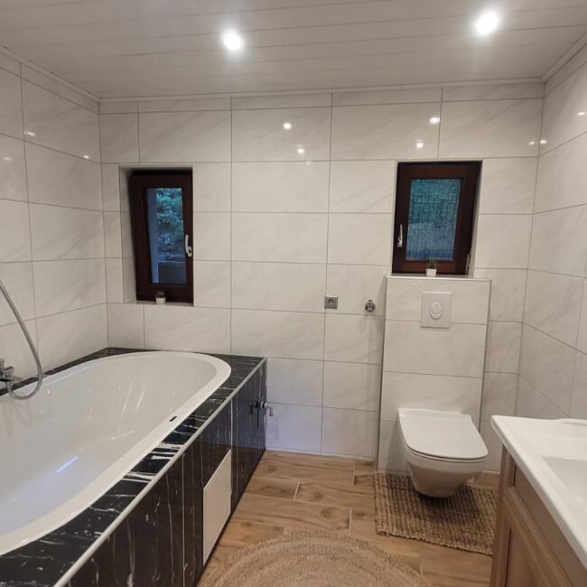 großes, helles Badezimmer mit einer großen Badewanne, Toilette, Waschbecken, 2 Fenstern und weißen sowie schwarzen Fließen.