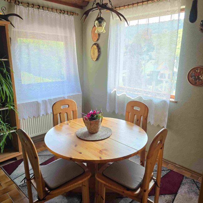 Esszimmer mit runden Esstisch aus Holz, um den 4 Holzstuhle stehen. Um den Tisch herum sind 2 große Fenster mit einem Blick auf den Pfälzerwald.