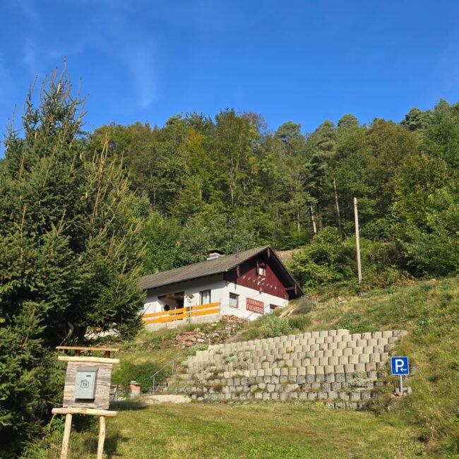 Ferienhaus Fuchsbau mit grünen Bäumen und blauen, wolkenlosen Himmel im Hintergrund. Im Vordergrund befindet sich ein Parkplatz und ein Holzschild mit einem Briefkasten.