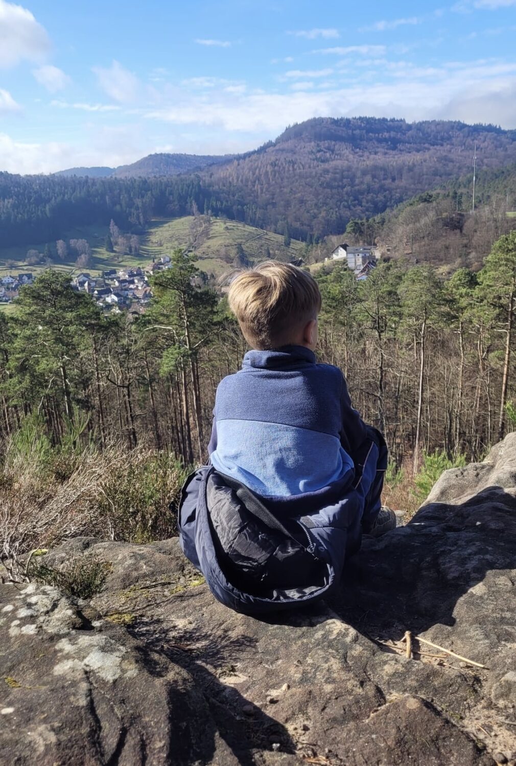 Kind sitzend auf einem Stein mit Blick auf die Stadt Igelbach. Im. Hintergrund sind Berge und blauber Himmel zu sehen.