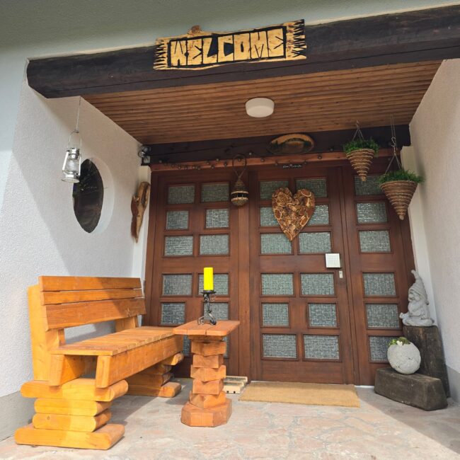 Eingangsbereich des Ferienhaus Fuchsbau mit einem "Welcome"-Schild, einer Holzbank und einer Holz-Tür mit Glasscheiben.
